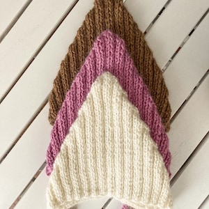 May include: Three knitted baby bonnets in different colours: brown, pink and white. The bonnets are stacked on top of each other, with the brown bonnet on top and the white bonnet at the bottom.