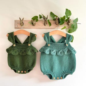 Könnte beinhalten: Zwei gestrickte Baby-Bodys in Grün und Türkis mit Rüschenbesatz und goldenen Knöpfen. Die Bodys hängen an Holzbügeln an einer weißen Wand.