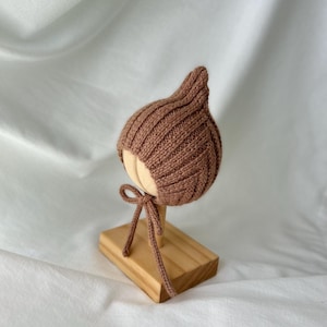May include: A brown knitted bonnet featuring a ribbed texture and a pointed top. The bonnet includes thin matching knitted ties that hang down from the chin area, designed to be tied securely under the chin for a snug fit around the head.