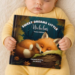Może przedstawiać: Książka dla dzieci zatytułowana "Sweet Dreams Little Nicholas - Forest Edition" z ilustracją śpiącego lisa. Książkę trzyma dziecko w ż&oacute;łtym pajacyku, leżące na kremowej, teksturowanej powierzchni.