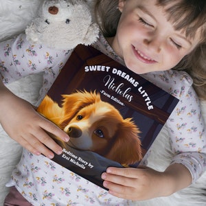 Puede incluir: Una ni&ntilde;a peque&ntilde;a sostiene un libro de cuentos para dormir titulado "Sweet Dreams Little Nicholas - Farm Edition." La portada del libro presenta un perro marr&oacute;n y blanco con el texto "Bedtime Story by Ersi Nicholls."