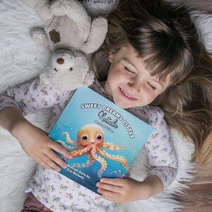 May include: A young girl is lying on a white fluffy blanket, holding a blue book with the title "Sweet Dreams Little Natalie - Ocean Edition". The book features an illustration of a pink and orange octopus with big eyes.