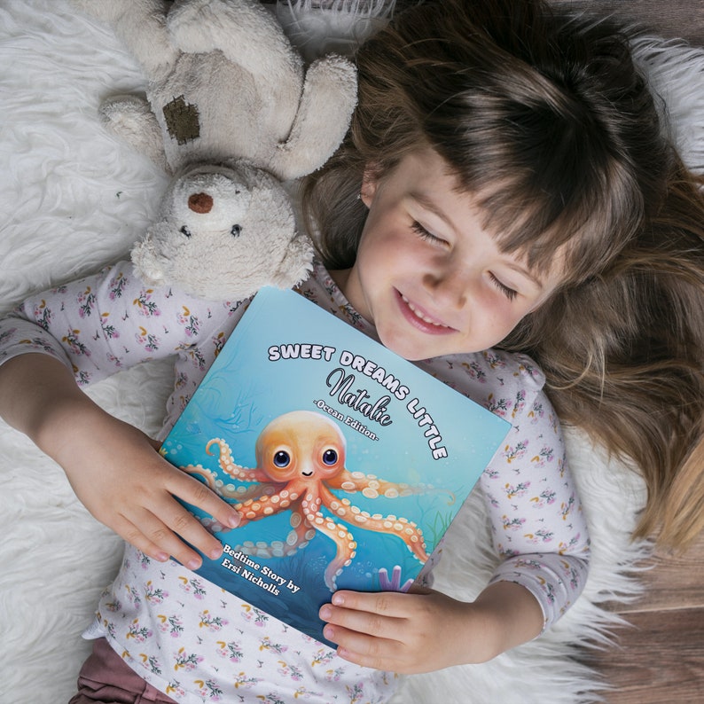 May include: A young girl is lying on a white fluffy blanket, holding a blue book with the title "Sweet Dreams Little Natalie - Ocean Edition". The book features an illustration of a pink and orange octopus with big eyes.