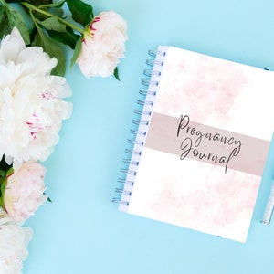 May include: A white spiral bound journal with a pink watercolour design and the text "Pregnancy Journal" on the cover. The journal is on a light blue background with white peonies.