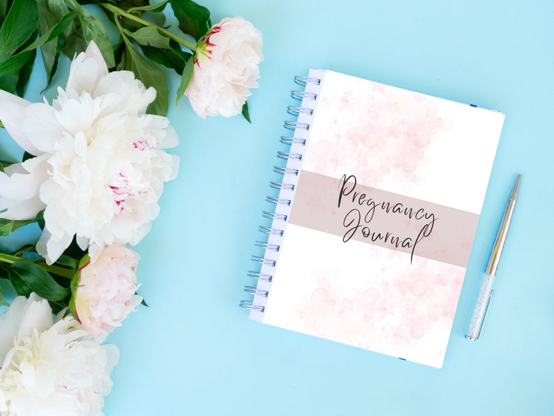 May include: A white spiral bound journal with a pink watercolour design and the text "Pregnancy Journal" on the cover. The journal is on a light blue background with white peonies.