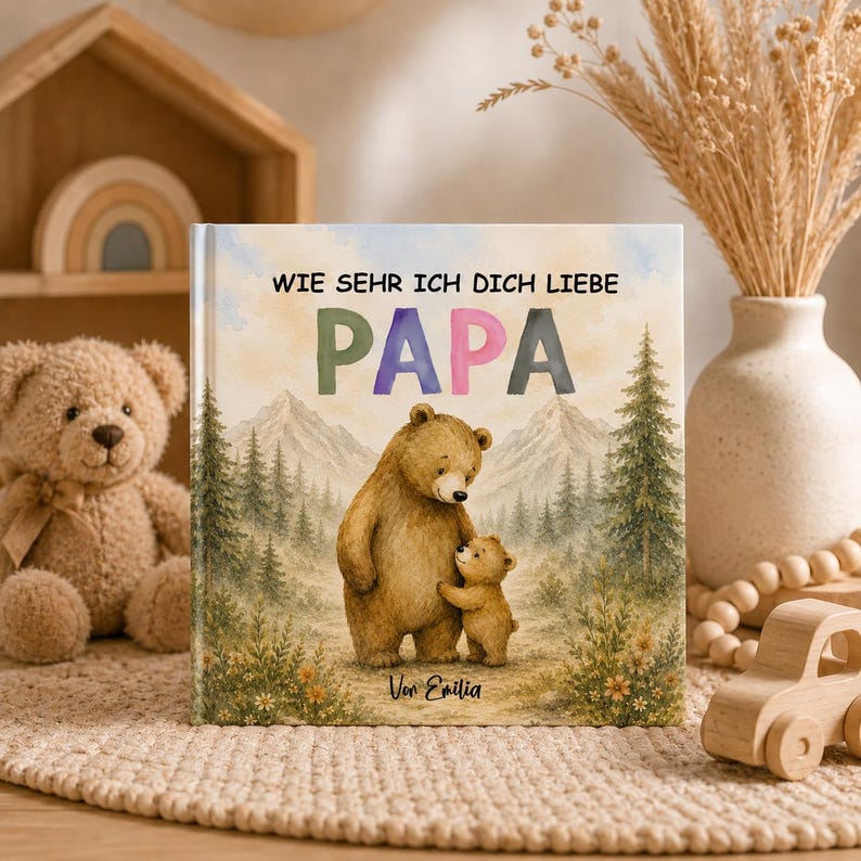 May include: A hardback book with the text "WIE SEHR ICH DICH LIEBE PAPA" and a watercolour illustration of a bear and cub in a forest setting. The book cover features a mountain backdrop and the name "Von Emilia".
