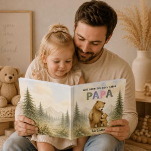 May include: A father and child are reading a book together. The book cover features a watercolor illustration of a bear and cub in a forest scene. The text on the cover reads "Wie Sehr Ich Dich Liebe Papa" and "Von Emilia".