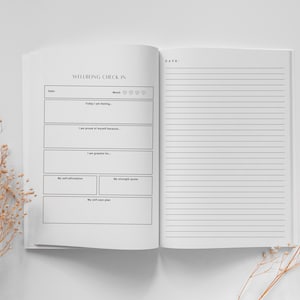 May include: A white journal with the title "Wellbeing Check In" and a section for daily self-reflection. The journal has sections for mood, self-affirmations, strength quotes, and a self-care plan. The right page is blank with lined paper.