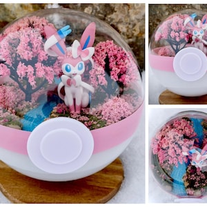 May include: A decorative terrarium inside a white and pink sphere, featuring a Sylveon figurine, pink cherry blossom trees, and a blue stream. The sphere is set on a wooden base, creating a miniature landscape.