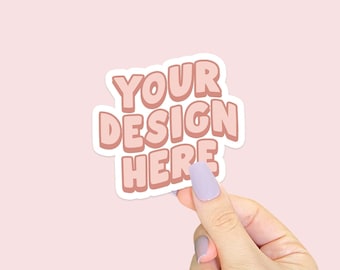 May include: A white sticker with pink text that reads "YOUR DESIGN HERE" being held by a hand with light purple nail polish.