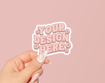 May include: A white sticker with pink text that says "YOUR DESIGN HERE" surrounded by small pink flowers. The sticker is being held by a hand with white nail polish.