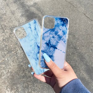Puede incluir: Dos fundas transparentes para tel&eacute;fono con dise&ntilde;os de m&aacute;rmol azul. Las fundas se sostienen en una mano con esmalte de u&ntilde;as azul claro.