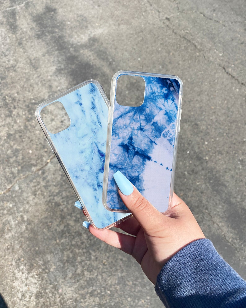 Puede incluir: Dos fundas transparentes para tel&eacute;fono con dise&ntilde;os de m&aacute;rmol azul. Las fundas se sostienen en una mano con esmalte de u&ntilde;as azul claro.