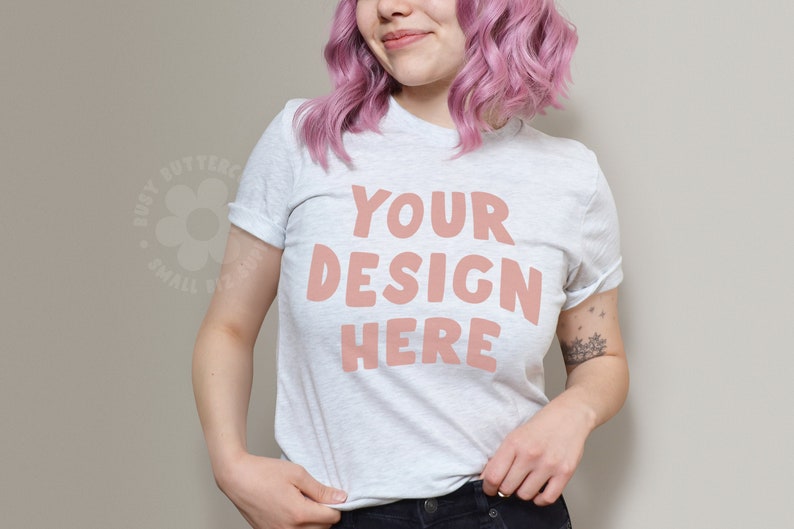May include: A white t-shirt with the text "YOUR DESIGN HERE" in pink lettering. The shirt is being worn by a person with pink hair.