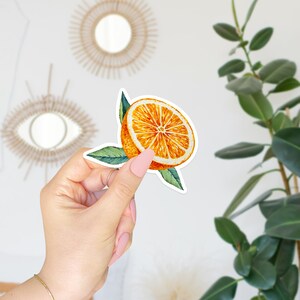 May include: A sticker of a half orange with green leaves. The orange is a vibrant yellow and orange color. The leaves are a deep green.