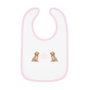 May include: White baby bib with pink trim and two embroidered golden retriever puppies wearing pink bows. The bib has the letters "LBK" embroidered in pink.