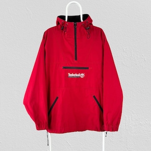 Puede incluir: Una chaqueta cortavientos roja de tipo pullover con cierre de media cremallera negro, capucha con cordones ajustables y un bolsillo tipo canguro frontal. El logotipo de Timberland Performance está bordado en blanco en la solapa del bolsillo.