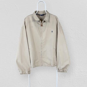 Peut inclure: Une veste zippée beige dotée d'un col classique, de manches longues à poignets élastiques et d'un petit logo bleu marine brodé sur la poitrine gauche. Le vêtement possède une fermeture éclair intégrale et deux poches latérales, sur un cintre.