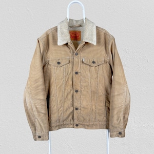 Puede incluir: Chaqueta de pana color canela con cuello de borreguillo color crema. La chaqueta tiene dos bolsillos en el pecho, dos bolsillos laterales y cierre de botones. Una etiqueta roja y blanca es visible en el pecho. La chaqueta está colgada de una percha blanca.