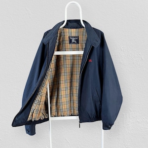 Op de afbeelding: Een marineblauw jack met rits en een voering in beige, zwart en rood ruitpatroon. Het binnenlabel bevat de tekst Burberrys met een ruiterlogo. Een klein rood geborduurd ruiterlogo is zichtbaar op de linkerborst van de marineblauwe stof.