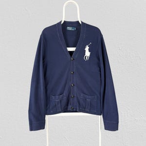 May include: A navy blue long-sleeve cardigan with a V-neckline and a button-front closure. The garment features a large white embroidered logo of a polo player on a horse on the left chest and two front patch pockets. A label reads Polo by Ralph Lauren.