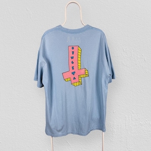 May include: Light blue t-shirt with a pink and yellow upside-down cross graphic on the back. The graphic has the word "OFWGKTA" printed vertically. The shirt is hanging on a white hanger against a white wall.