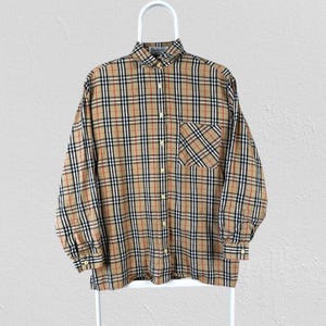 May include: A long-sleeved shirt with a beige, black, red, and white plaid pattern. The shirt has a button-down front, a chest pocket, and buttoned cuffs. It is displayed on a white hanger. The shirt is a classic design.