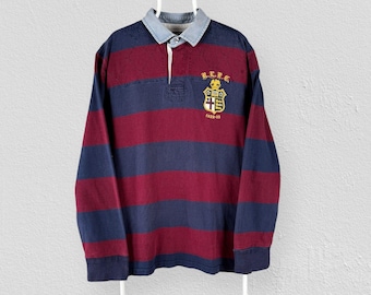 May include: A long-sleeve rugby shirt featuring horizontal stripes in dark navy blue and deep burgundy. The shirt has a light blue collar and a button placket. An embroidered crest patch in yellow, white, and red with the text B.L.P.C. 1922-33.