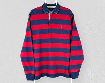 May include: A long-sleeve rugby shirt featuring horizontal red and navy blue stripes. The shirt has a dark blue denim collar, a button-up placket, and a small blue embroidered logo on the left chest. The cuffs are solid navy blue.