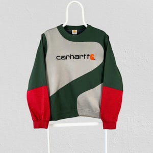 Peut inclure: Un sweat-shirt à col rond et manches longues présentant un design color-block en vert foncé, gris clair et rouge. L'avant affiche le texte "carhartt" en lettres noires avec un logo "c" orange. Le vêtement est présenté sur un cintre blanc.