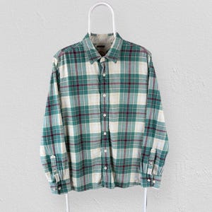 Peut inclure: Une chemise à carreaux à manches longues dans les tons de vert, blanc et bordeaux est suspendue à un cintre blanc. La chemise présente une fermeture boutonnée, un col et des poignets boutonnés. Le motif à carreaux est uniformément espacé.