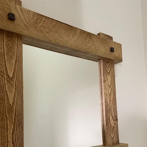 May include: A rustic wooden mirror with a rectangular frame. The frame is made of thick, brown wood with a natural finish. The mirror is surrounded by a simple, rectangular frame. There are four black metal accents on the frame.