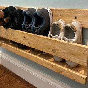 May include: A wall-mounted shoe rack made of light-coloured wood, holding several pairs of shoes. The rack is fixed to a light blue wall. Various shoes are stored on the three shelves, including trainers and casual footwear.