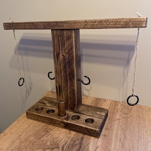 May include: A wooden ring toss game with a dark stain. It has a horizontal bar, a vertical support, and a base with holes. Black metal hooks and rings are attached with twine. The game is set on a wooden surface, ideal for indoor or outdoor play.