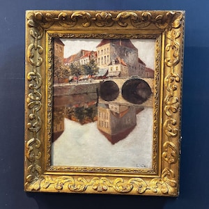 Peut inclure: Peinture à l'huile d'un paysage urbain européen avec un pont sur une rivière, encadrée dans un cadre doré orné. La peinture représente des bâtiments avec des toits en tuiles rouges et leurs reflets dans l'eau. Le cadre mesure environ 46 x 61 cm.