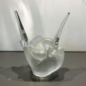 Peut inclure: Une sculpture en verre transparent représentant un oiseau aux ailes déployées. L'oiseau est niché dans une base en verre dépoli, avec deux longs éléments en verre pointus s'étendant vers le haut. La sculpture est exposée sur une surface grise.