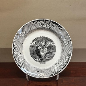 May include: A black and white decorative plate with a detailed border design. The centre of the plate features a detailed illustration of a person sitting under a tree. The plate is displayed on a clear stand.