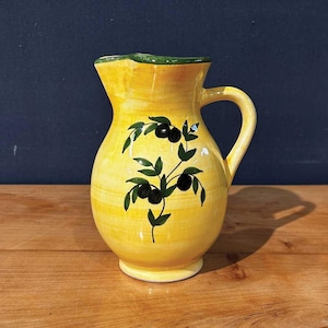 Peut inclure: Une cruche en céramique jaune avec un bord et une anse verts. La cruche est décorée d'un motif de branche d'olivier peint avec des olives noires et des feuilles vertes. La cruche est posée sur une surface en bois.