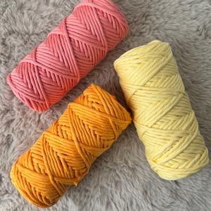 Op de afbeelding: Drie spoelen katoenen touw in verschillende kleuren: roze, oranje en geel. Het touw is dik en heeft een gevlochten textuur.