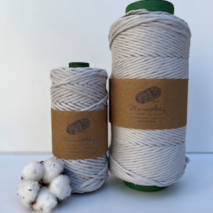 Peut inclure: Deux bobines de fil macramé gris clair avec des étiquettes marron portant le texte "Macram Akkiz". Une branche de coton est au premier plan. Les bobines sont sur une surface blanche.