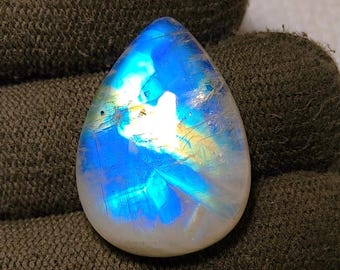 May include: A pear-shaped, polished moonstone gemstone. The stone displays a vibrant play of color, with iridescent blues, whites, and yellows. The gemstone is set against a dark green background.