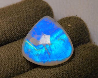 May include: A triangular, iridescent gemstone with a blue and white color scheme. The stone has a polished surface and displays a play of light, creating a shimmering effect. The gemstone is held in a dark green glove.