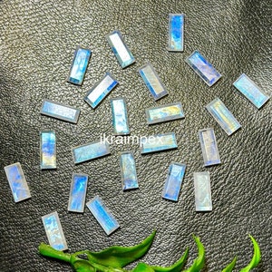 May include: A collection of 18 iridescent blue-green rectangular gemstone cabochons, scattered on a black leather surface. The stones have a shimmering, opalescent quality.