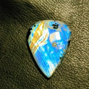 May include: A blue and gold labradorite gemstone with a unique, iridescent pattern. The gemstone is cut in a teardrop shape and has a smooth, polished surface.