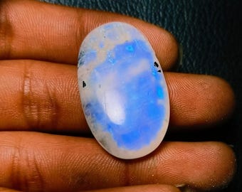 Può includere: Un grande cabochon di pietra di luna di forma ovale con una base bianca lattiginosa e riflessi iridescenti blu e bianchi. La pietra è liscia e lucidata.