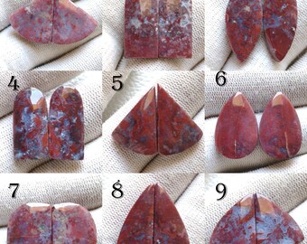 May include: Nine pairs of polished gemstone cabochons in various shapes, including teardrop, triangular, and rectangular. The stones display a mix of red, burgundy, and blue-grey hues, with natural patterns. The cabochons are likely for jewelry making.