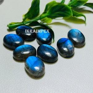 May include: Eight oval-shaped labradorite gemstones with iridescent blue and black colors. The stones are arranged in a semi-circle on a white surface. The text "IKRAIMPEX" is visible in the image.