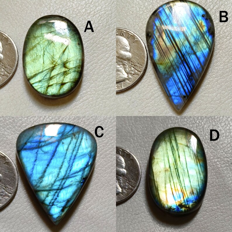 Peut inclure: Quatre cabochons de labradorite en diff&eacute;rentes formes et tailles. Les pierres sont iris&eacute;es avec des nuances de bleu, de vert et d'or. Les pierres sont pr&eacute;sent&eacute;es sur une surface blanche avec un quart de dollar am&eacute;ricain pour l'&eacute;chelle.