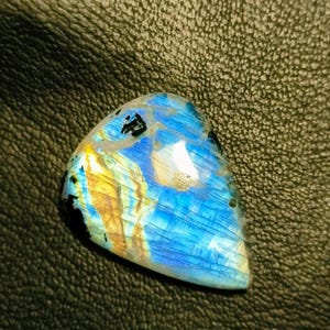 May include: A blue, gold, and black labradorite gemstone cabochon with iridescent shimmer. The gemstone is triangular in shape and has a smooth, polished surface.