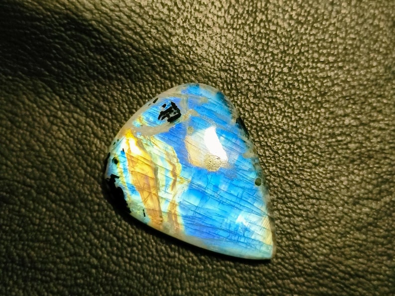 May include: A blue, gold, and black labradorite gemstone cabochon with iridescent shimmer. The gemstone is triangular in shape and has a smooth, polished surface.